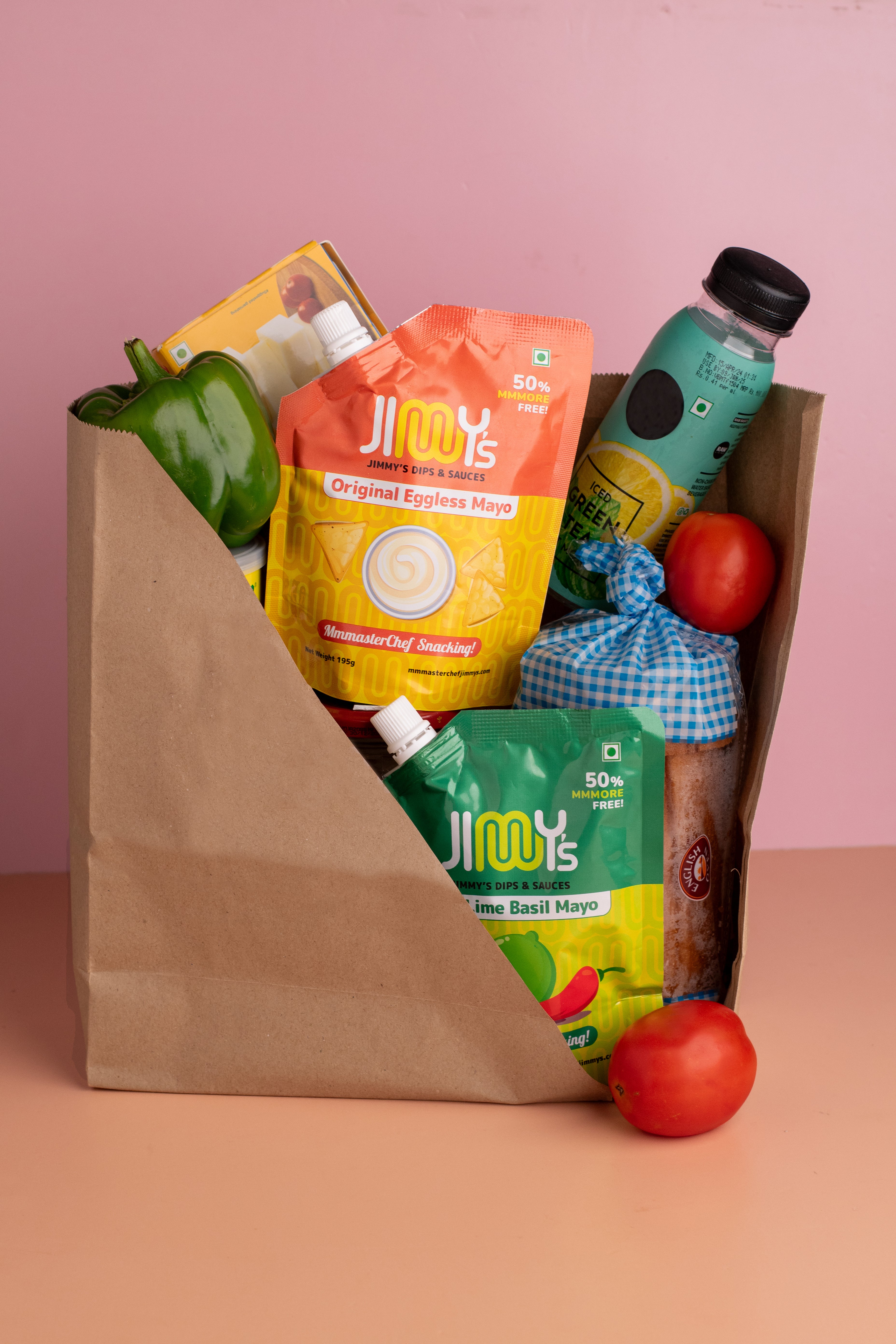 Paper bag filled with groceries including a bottle of Jimmy's Original Express Noodle, a green bell pepper, and tomatoes on a pink background.